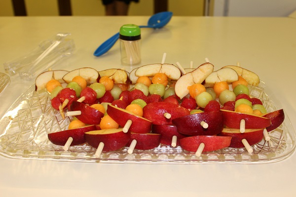 frigarui de fructe puse pe o tava transparenta, frigaruile continand piersici sau nectarine, trei feluri de pepene si mar, tava cu frigarui aflandu-se langa un recipient cu scobituri si langa o lingura albastra de plastic, pe o masa