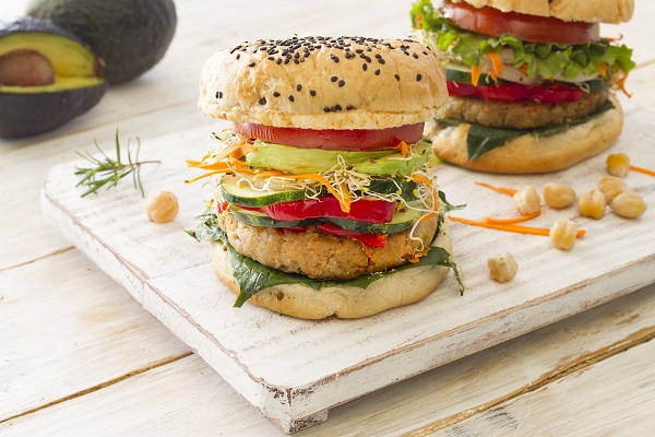 blat de lemn alb pe care se afla doi hamburgeri in chifle cu seminte, burgerii fiind vegetarieni, din naut, avand si salata, rosii, avocado si putin morcov ras, pe blatul alb de lemn fiind si cateva boabe de naut