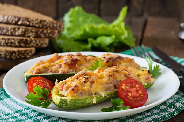 barcute de zucchini la cuptor, cu umplutura de carne, cu crusta de branza, puse pe o farfurie alba decorata cu rosii si cu bucati de patrunjel, farfuria fiind asezata pe un servet cu carouri verzi si albe, langa farfurie aflandu-se tacamuri, foi de salata si cateva felii de paine integrala cu susan