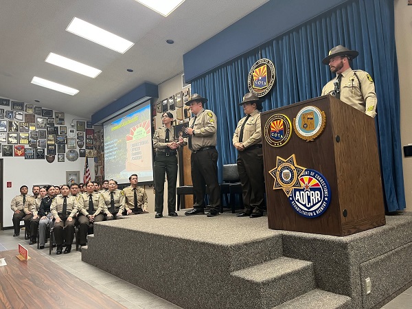 politisti de la Biroul Serifului din Gila Arizona, in uniforme, unul dintre ei stand la pupitru si facand un anunt celor care se afla intr-o sala stand pe scaune