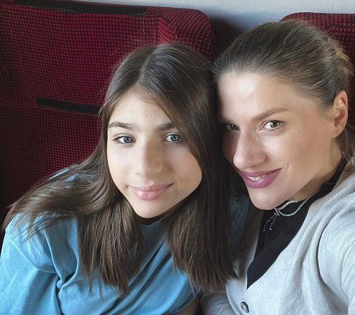 Tily Niculae vesela, cu parul strans in coada, imbracata cu bluza neagra si pulover gri deschis, in timp ce sta canapeaua visinie dintr-un tren alaturi de fiica ei, Sofia, imbracata cu bluza bleu