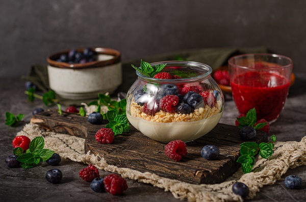 desert panna cotta, cu cereale de ovaz si fructe de padure proaspete si frunze de menta deasupra, pus intr-un recipient de sticla, aflandu-se pe un tocator de lemn inchis la culoare, cu zmeura si afine imprastiate, alaturi de care se afla un recipient de sticla cu gem de fructe de padure si castronele cu zmeura si cu afine