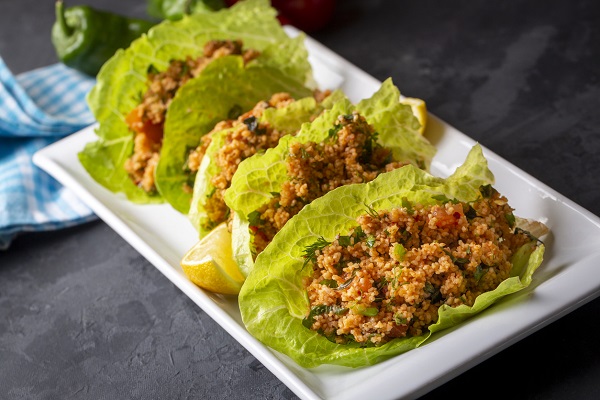 salata de bulgur cu legume, pusa pe frunze de salata, alaturi de o bucata de lamaie,asezate pe o tava alba, pusa pe o suprafata gri pe care se afla un servet de bucatarie alb cu bleu