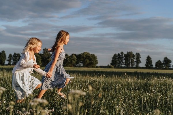 doua fetite blonde imbracate in rochite, una dintre fetite avand rochita alba si puloveras si cealalta rochita cu dungi albe si negre, tinandu-se de mana in timp ce aleaga pe un camp intr-o zi de vara