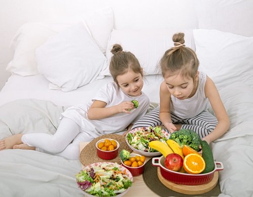 doua fetite imbracata cu pijamale, cu parul strans in coc,stand intr-un pat cu lenjerie alba, avand in fata lor castroane cu salate, cu rosii cherry portocalii si cu alte fructe si legume proaspete, una dintre fetite tinand in mana un buchetel de broccoli