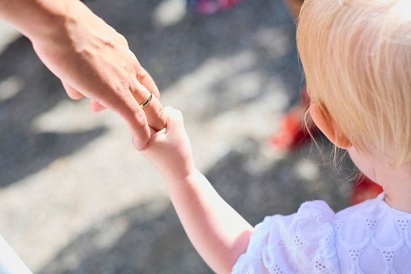 fetita blonda, imbracata in alb, tinuta de manuta de o femeie care poarta verigheta, amandoua aflandu-se afara