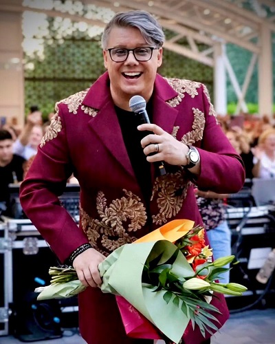Paul Surugiu Fuego vesel, imbracat cu sacou visiniu de catifea cu auriu, in timp ce tine cu o mana un microfon si cu celalalta un buchet de flori, in timp ce se afla pe scena, la un concert, avand in spatele sau publicul