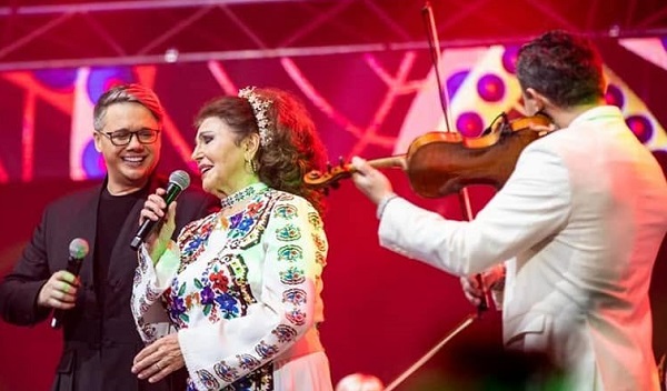 Fuego purtand ochelari, imbracat cu costum negru, in timp ce tine in mana un microfon si o priveste zambind pe Irina Loghin, imbracata in costum popular, in timp ce se afla amandoi pe o scena, langa ei fiind un violonist imbracat in alb