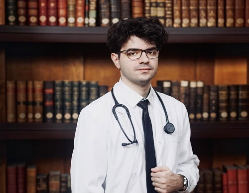 Calin Vamesu, tanarul care a iesit sef de promotie la Facultatea de Medicina din Iasi, purtand ochelari, imbracat cu halat alb si cu stetoscop, in fata unei biblioteci pline cu carti