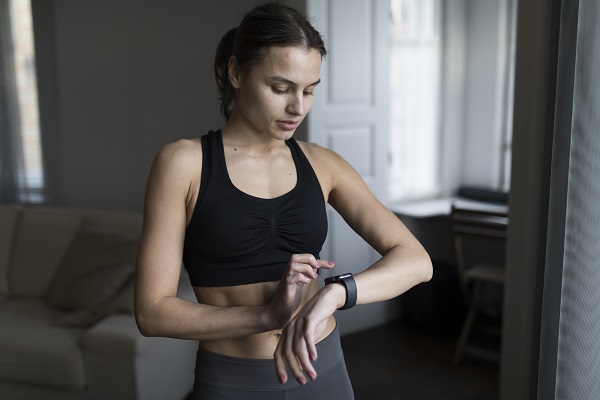 femeie tanara, cu parul prins in coada, imbracata cu bustiere neagra si colanti, in timp ce se uita la un smartwatch cand se afla intr-o camera a casei