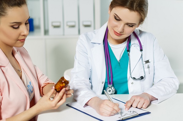 doctorita tanara, zambitoare,cu vesta turcoaz, bluza aba si halat, cu stetoscop violet, in timp ce sta la birou in cabinet si scrie o recomandare sau o reteta pentru pacienta ei, imbracata in roz, care tine in mana un flacon de medicamente si se uita la acesta
