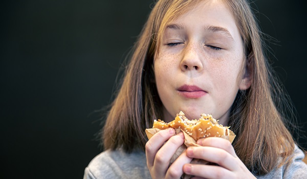 adolescenta care tine ochii inchisi in timp ce savureaza un hamburger pe care il tine in mana