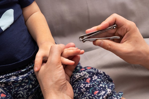 persoana adulta care taie cu un cleste unghiile unui copil imbracat cu tricou bleumarin si cu pantalonasi bleumarin cu imprimeu floral, care sta asezat pe o canapea gri