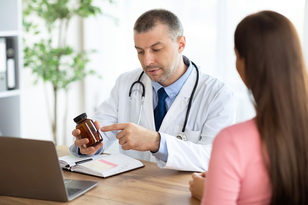 femeie tanara cu parul lung, imbracata in roz, stand pe scaun si uitandu-se la medicul aflat la birou, care ii arata un recipient cu suplimente alimentare sau cu medicamente