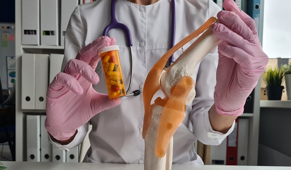 doctorita imbracata cu halat alb, purtand manusi de unica folosinta roz, avand stetoscop la gat, cu o mana tinand un flacon cu medicamente si cu cealalta un mulaj de os cu tendoane si ligamente, in timp ce in spatele ei se afla un biblioraft din cabinet