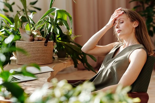 femeie imbracata cu rochie oliv fara maneci, ducandu-si mana la frunte din pricina unei dureri de cap pe care o are in timp ce sta sa lucreze la un birou, avand laptopul in fata si fiind inconjurata de plante