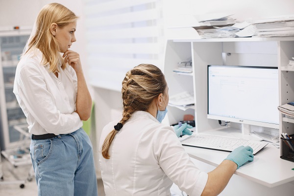 femeie tanara,blonda, imbracata cu camasa alba si blugi, tinandu-si mana la barbie si stand langa un cadru medical cu parul prins in coada impletita, care sta pe scaun la computer la un birou alb intr-un cabinet medical