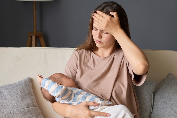 mama tanara, imbracata cu tricou maroniu, ducandu-si mana la frunte intrucat nu se simte bine in timp ce sta pe o canapea bej cu perne decorative gri pentru a-si alapta bebelusul, imbracat cu bluza alba cu dungi bleu si cu pantoni albi