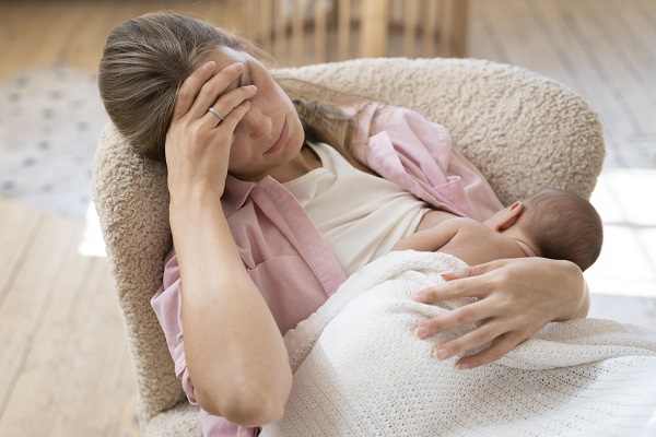 mama imbracata cu maieu alb si camasa roz, ducandu-si mana la frunte si avand ochii inchisi in timp ce sta pe un fotoliu bej si isi tine in brate bebelusul, invelit pe jumatate de corp cu o paturica alba, ca sa-l alapteze