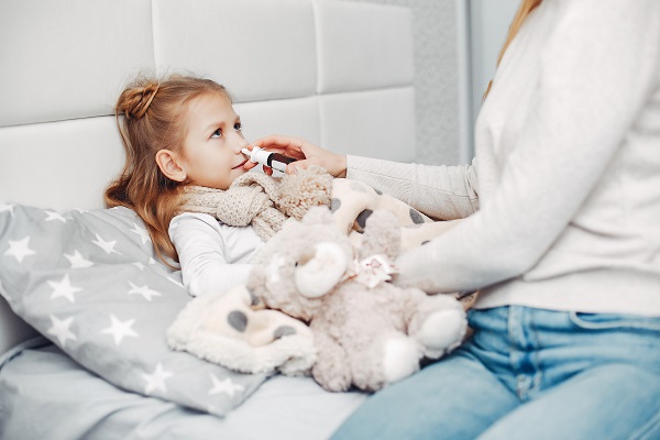 fetita imbracata cu pijama alba, cu fular gri la gat, care sta rezemata de o perna gri cu stelute albe, in sezut, in pat, tinand langa ea un ursulet de plus, in timp ce mama ei, imbracata cu blugi si pulover incearca sa ii pulverizeze un spray nazal
