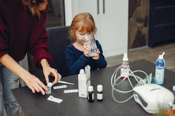 fetita cu parul roscat, imbracata cu bluza de casa bleumarin, tinand in mana un pahar cu apa, in timp ce sta la o masa pe care se afla spray nazal, nebulizator, termometru si diverse medicamente, in timp ce mama ei, imbracata cu pulover visiuniu si blugi, toarna ceva intr-un mic recipient