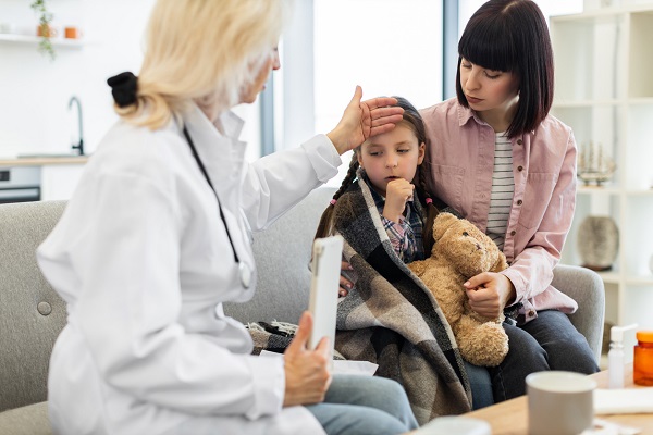 mama imbracata cu camasa roz, stand pe canapea langa fetita ei cu codite, invelita cu o patura pe umeri, care tuseste, in timp ce o doctorita blonda sta langa ea si ii pune mana pe frunte sa-i verifice temperatura