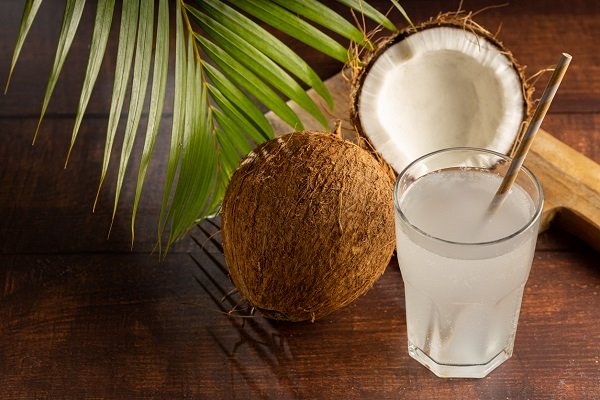nuca de cocos, frunza de cocotier si pahar de sticla cu apa de cocos si pai, toate asezate pe o suprafata de lemn