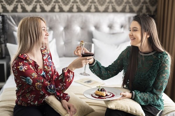 doua femei vesele, una dintre ele imbracata cu bluza visinie cu flori si cealalta cu bluza verde de dantela, stand pe un pat, fiecare dintre ele avand cate o pernuta crem pe genunchi, una dintre ele tinand in mana un pahar de vin rosu, iar cealalta o furculita cu o bucatica de prajitura si o farfurie cu prajitura ornata cu fructe