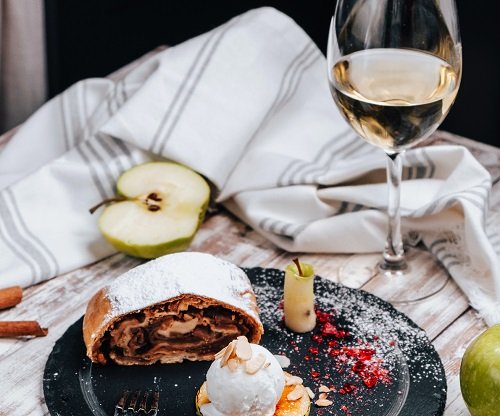 pahar de vin alb, jumatate de mar verde si farfurie neagra pe care se afla o bucata de strudel cu mere pudrat cu zahar si o prajitura cu inghetata pe deasupra, langa farfurie fiind un baton de scortisoara, un prosop de bucatarie cu alb si gri si alt mar verde, toate asezate pe o masa de lemn albicioasa