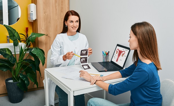doctorita tanara, zambitoare, care tine in mana imaginile de la o ecografie si ii da explicatii pacientei sale, imbracata cu bluza albastra, in timp ce amandoua stau pe scaun la biroul doctoritei in cabinet, pe birou fiind si un laptop care arata o imagine cu aparatul reproducator feminin