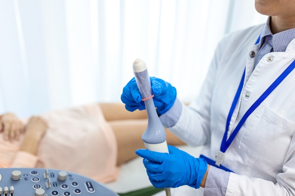 cadru medical in halat, cu manusi albastre, care se pregateste sa utilizeze un dispozitiv pentru realizarea unei ecografii transvaginale pentru o pacienta in camasa roz, care sta intinsa pe un pat