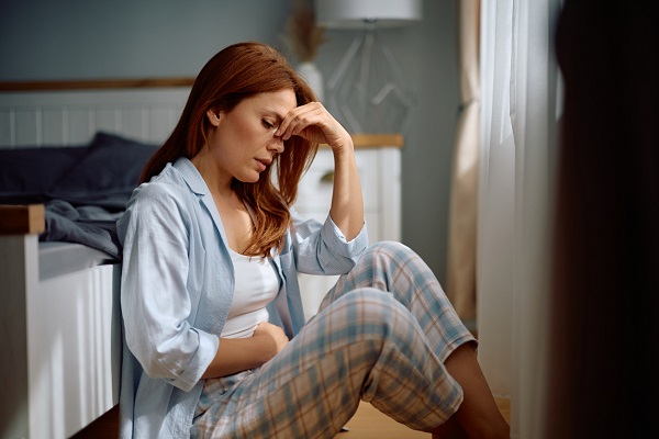 femeie cu parul roscat, imbracata in pijama cu bluza bleu si pantaloni in carouri, stand pe podea, langa pat, intrucat nu se simte bine, ducandu-si o mana la frunte si una in zona abdominala