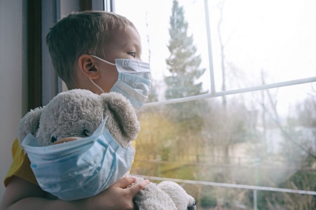 copil cu masca de protectie uitandu-se pe geam, cu urs de plus in mana