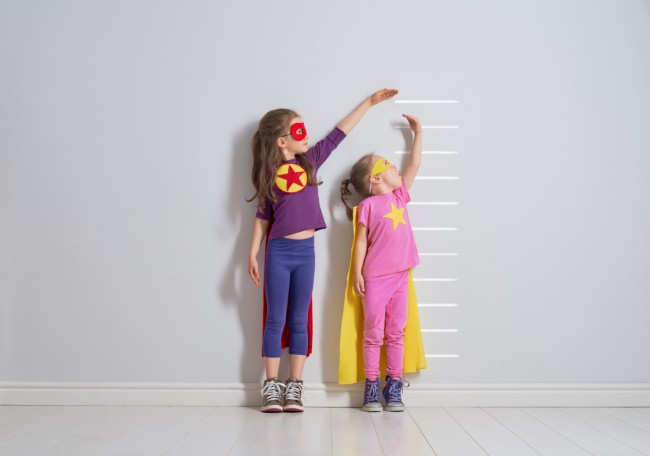 doua fetite imbracate in costume de supereroi care isi masoara inaltimea langa un perete
