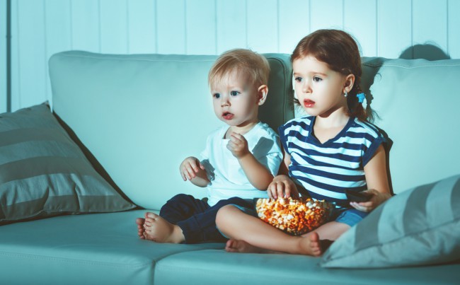 doi copii, baiat si fetita, care stau pe canapea si se uita la televizor in timp ce mananca popcorn