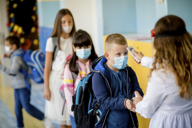 mai multi elevi cu masca ce stau la rand pentru a li se lua temperatura de catre profesoara inainte de a intra in clasa