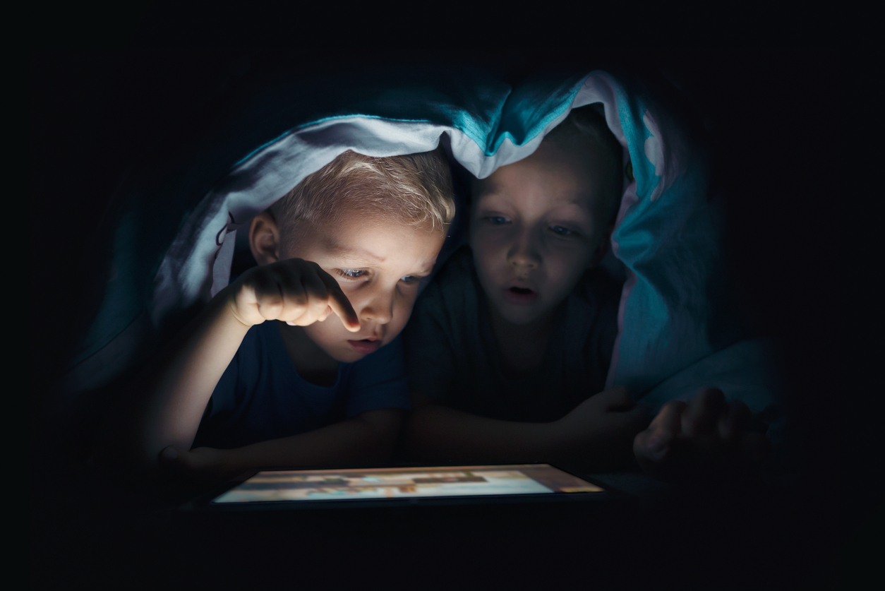 doi copii care se uita pe tableta sub patura, in intuneric