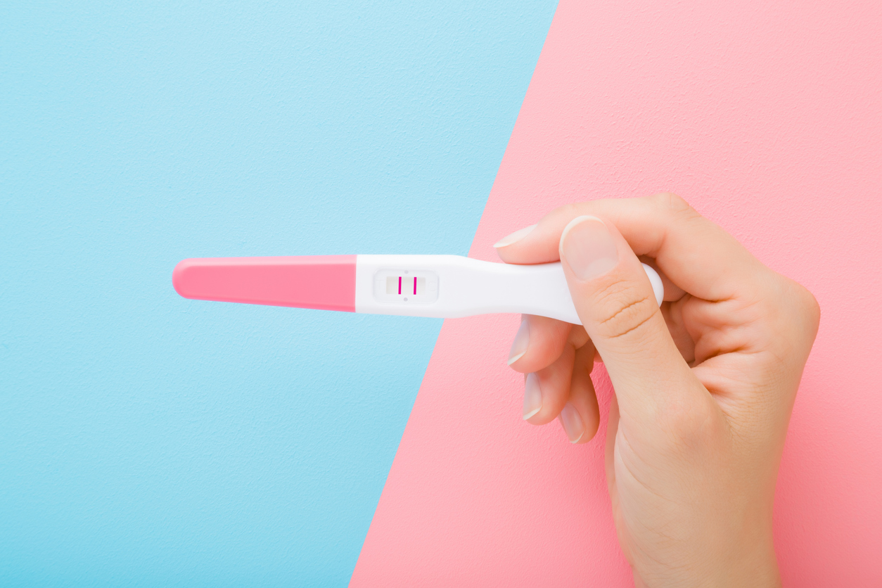 femeie care tine in mana un test de sarcina pozitiv, cu doua liniute, pe fundal roz cu albastru