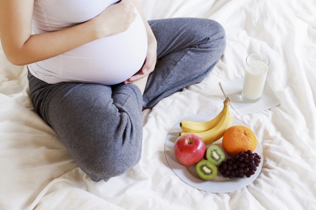 femeie insarcinata care sta pe pat cu un platou de fructe si un pahar cu lapte in fata