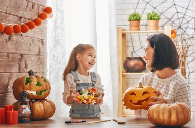 fetita care tine in mana un bol cu dulciuri si zambeste la mama ei, care tine in mana un dovleac sculptat