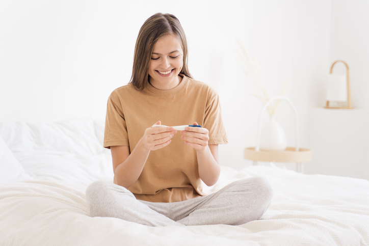 femeie fericita care sta pe pat si tine in maini un test de sarcina