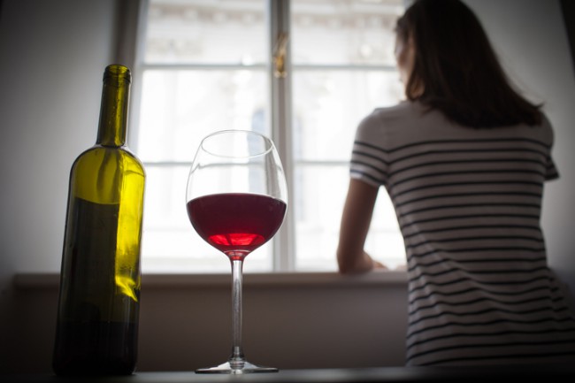 femeie care sta cu spatele si se uita pe fereastra; langa ea sunt o sticla de vin si un pahar