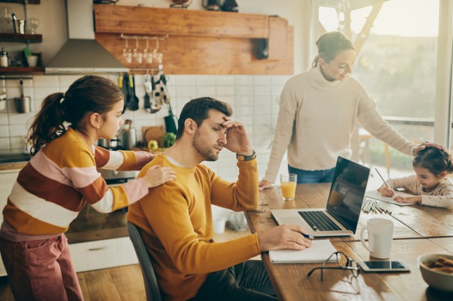 Tată extenuat care lucrează la laptop în timp ce se află acasă cu familia sa