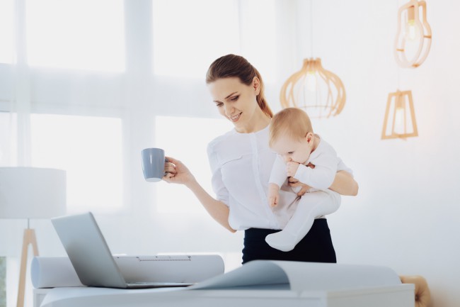 mama care lucreaza si tine in mana dreapta o cana iar in cealalta un bebelus si se uita in ecranul laptopului