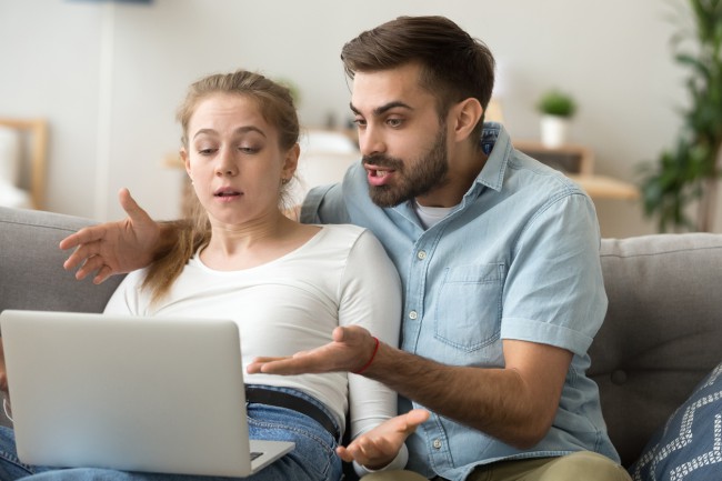 barbat si femeie care stau pe canapea si privesc intr-un laptop furiosi