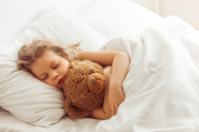 fetita care doarme in pat cu un ursulet de plus in brate