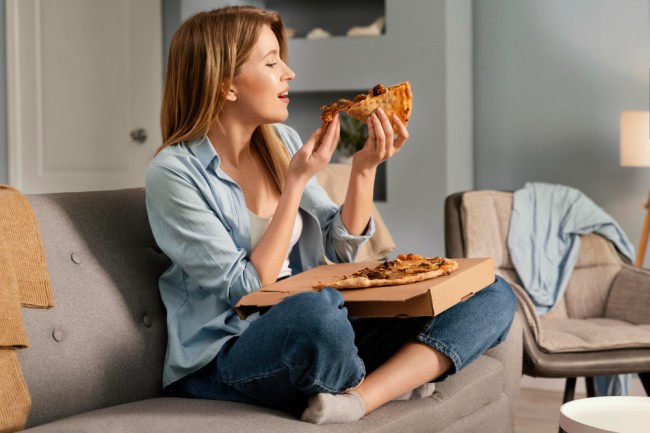 femeie care sta pe canapea si mananca o pizza