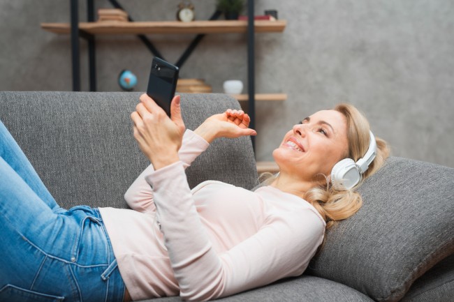 femeie care sta pe canapea cu un telefon in mana si asculta muzica la casti
