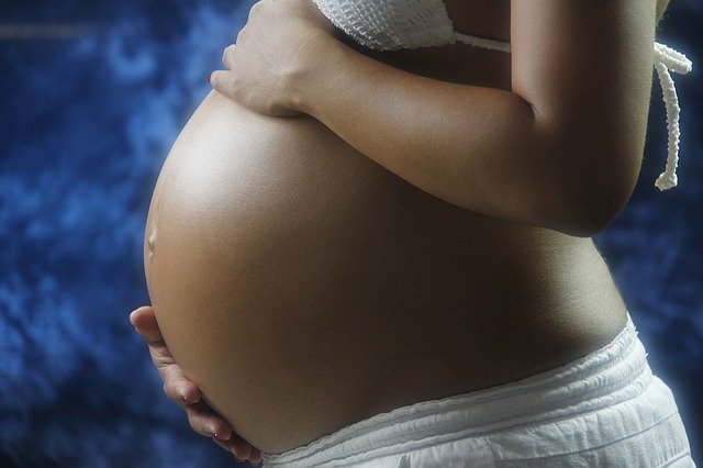 burta goala de femeie gravida asezata intr-o parte cu mainile pe abodmen imbracata in alb