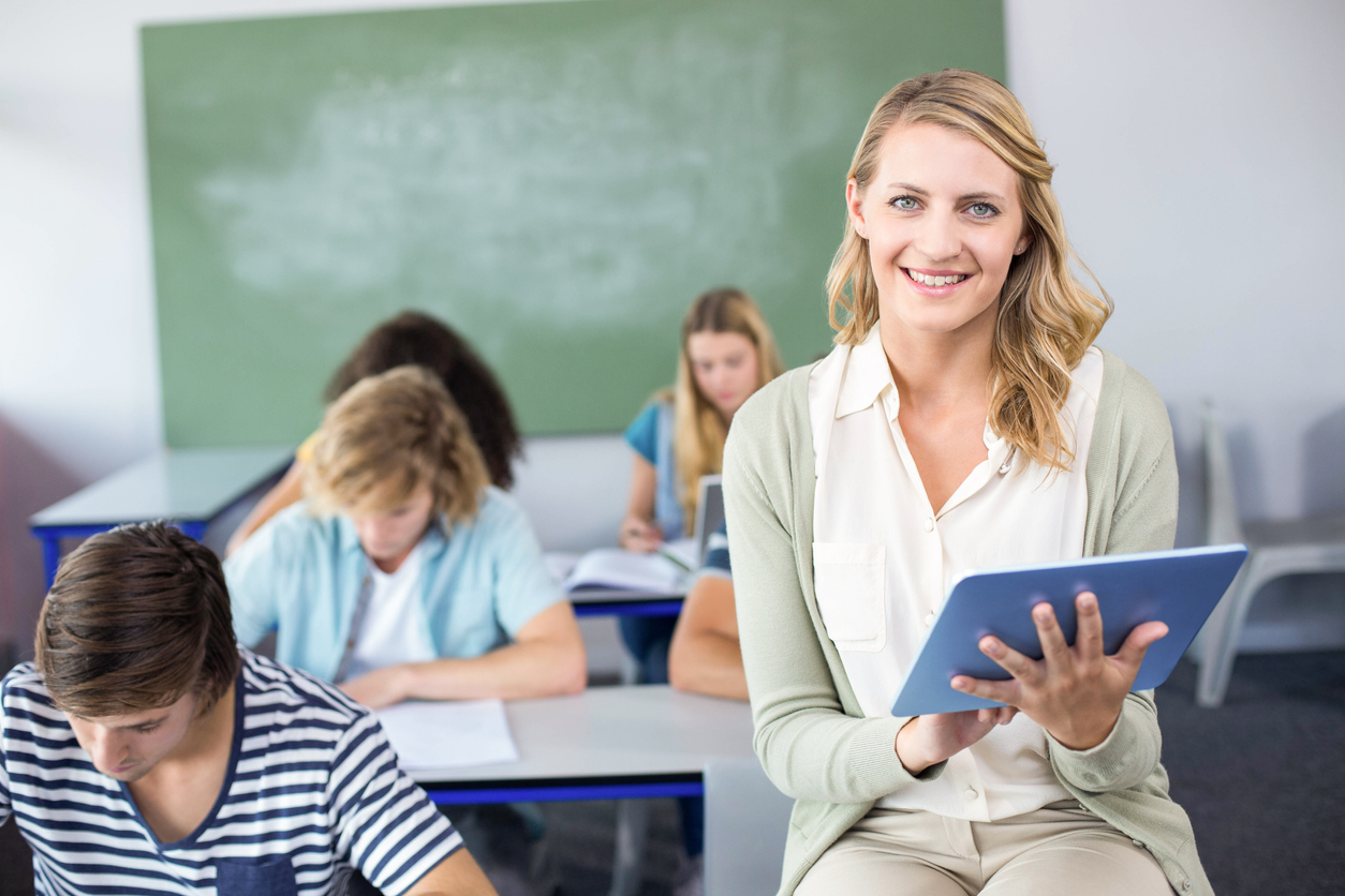 profesor de liceu cu tableat in mana cu banci cu copii in fundal